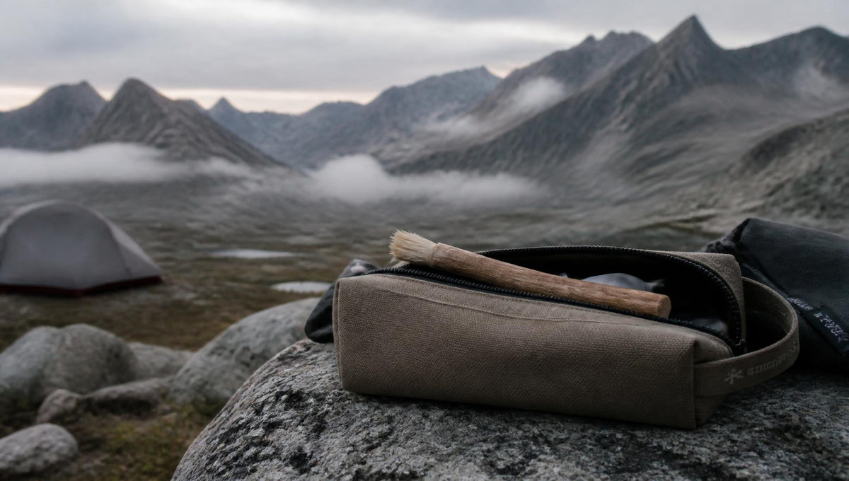 A brown canvas zippered pouch with a wooden-handled brush resting inside, placed on a large rock in a mountain landscape with misty peaks and a tent in the background.